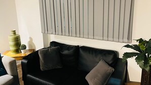 A black leather sofa with two cushions, a small wooden side table with a green vase and a potted plant, and a window with vertical blinds. - 5 Minute Walk to St Paul s Cathedral (London)