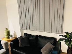 A black leather sofa with two cushions, a small wooden side table with a green vase and a potted plant, and a window with vertical blinds. - 5 Minute Walk to St Paul s Cathedral (London)