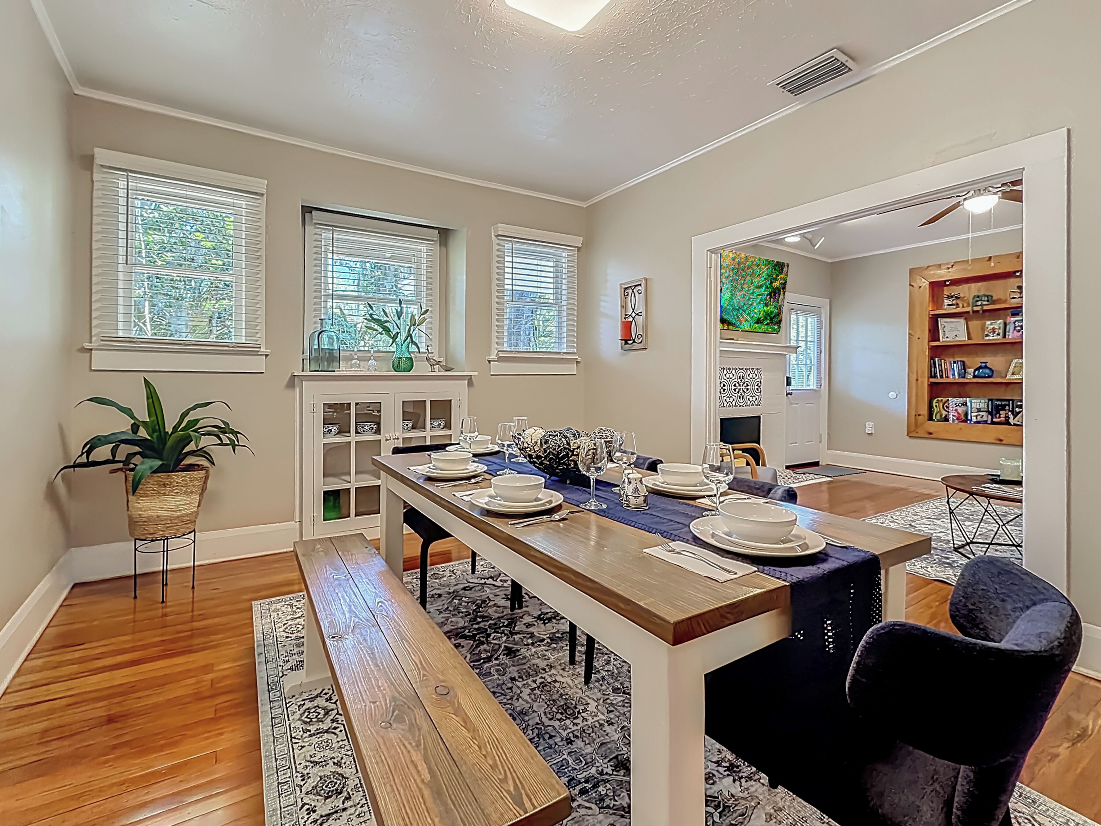 A dining area with a long wooden table set for six, flanked by two windows with blinds, a potted plant, and a bookshelf.
