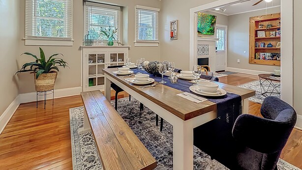 A dining area with a long wooden table set for six, flanked by two windows with blinds, a potted plant, and a bookshelf. - The Peacock House - Vintage Downtown Charm (Ocala)