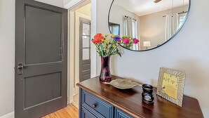 A hallway with a dark door, a mirror, a vase with flowers, a decorative plate, and a framed picture on a wooden dresser. - The Peacock House - Vintage Downtown Charm (Ocala)