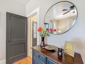 A hallway with a dark door, a mirror, a vase with flowers, a decorative plate, and a framed picture on a wooden dresser. - The Peacock House - Vintage Downtown Charm (Ocala)