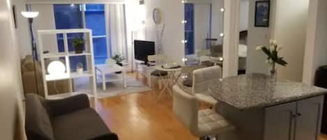 Una sala moderna con un sofá, un comedor con mesa y sillas, una cocina con barra, y un balcón con vistas.