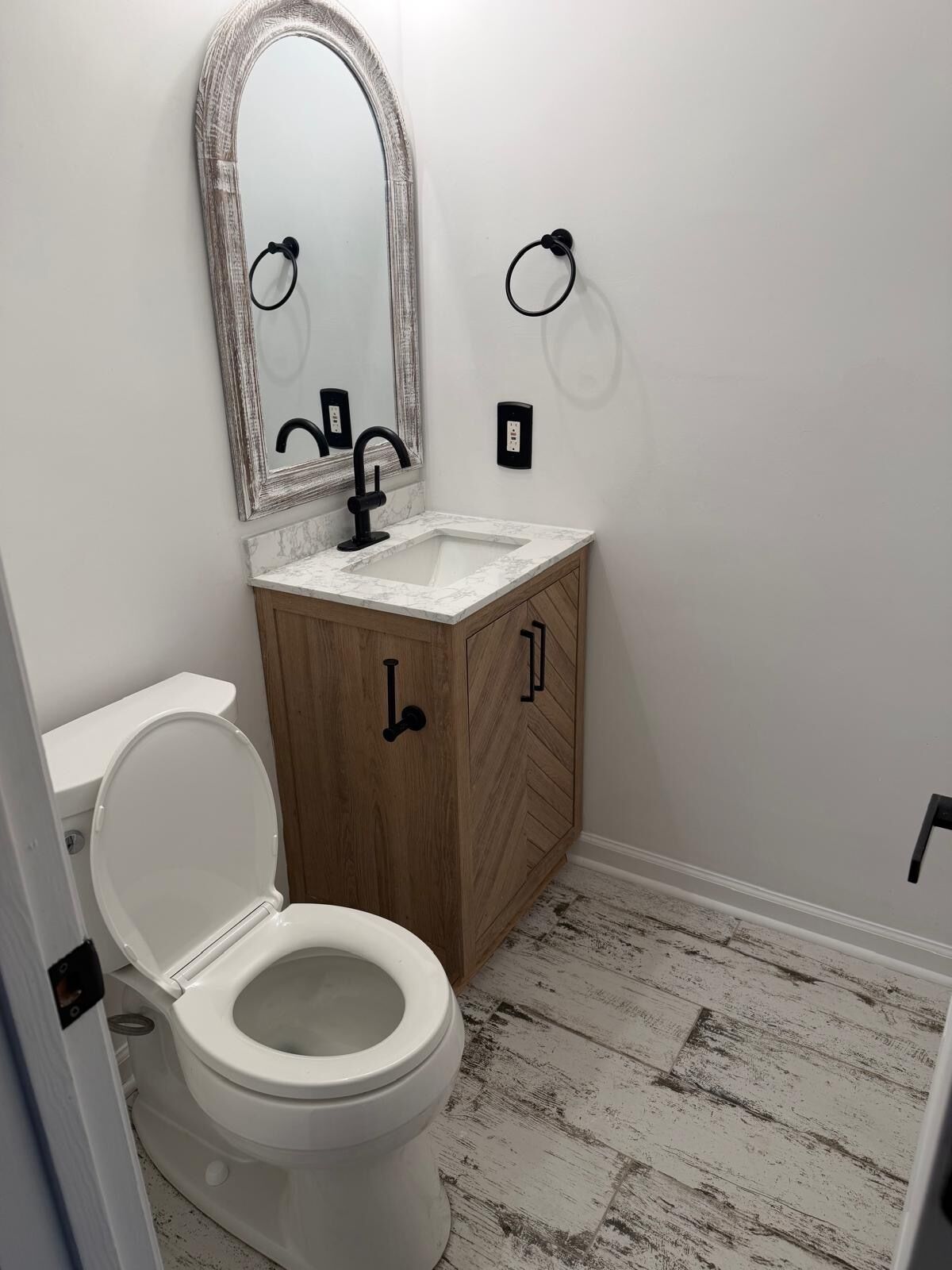 A bathroom with a white toilet, a wooden vanity with a marble countertop, a large mirror, and a black faucet.