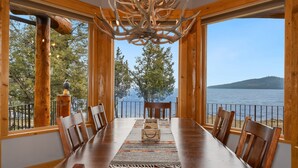 A dining area with a wooden table, chairs, a chandelier, and a view of a lake through large windows.