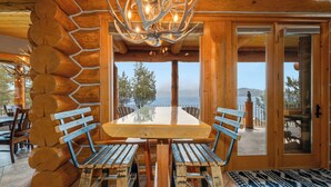 A wooden interior with a large table and benches, a chandelier, and a view through the glass doors.