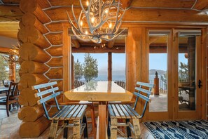 Un intérieur en bois avec une grande table et des bancs, un lustre et une vue à travers les portes vitrées.