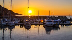 Un port de plaisance où sont amarrés de nombreux bateaux, un coucher de soleil en arrière-plan et une côte rocheuse.