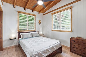 Ein Schlafzimmer mit einem Bett, zwei Nachttischen, einer Holzkommode und großen Fenstern mit Jalousien.