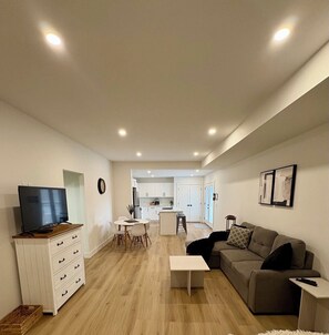 A modern living room with a TV, white furniture, a sofa, and a kitchen in the background.