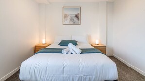 Un lit bien fait, avec du linge de lit blanc, deux lampes de chevet, un tableau encadré au mur et une petite table de chevet en bois.
