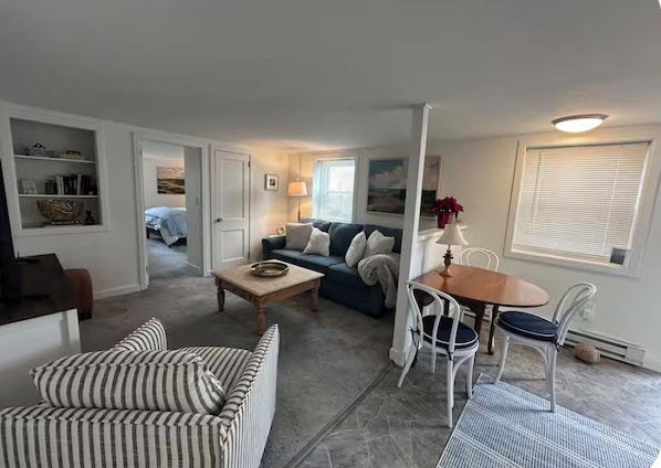 A living room with a sofa, a coffee table, a dining table with chairs, a bookshelf, and a door leading to another room.