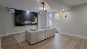 A living room with a large flat-screen TV mounted on the wall, a beige sectional sofa, wooden flooring, and a ceiling-mounted projector.