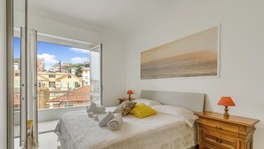Een slaapkamer met een bed, een nachtkastje, een raam met uitzicht op gebouwen en een schilderij aan de muur.