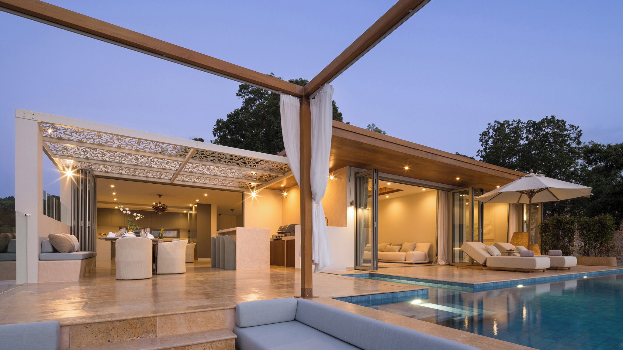 Un espace piscine extérieur moderne doté d’un coin salon couvert, d’un barbecue et d’un salon fermé par des baies vitrées.