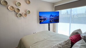 Une chambre à coucher avec un lit, un téléviseur fixé au mur et une fenêtre donnant sur des immeubles.