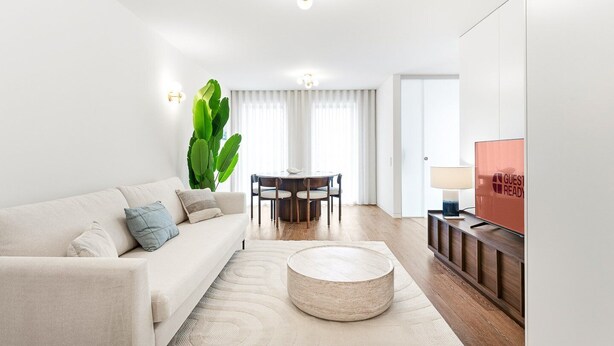 En moderne stue med sofa, rundt sofabord, spisebord med stole, en stor plante og et fjernsyn.