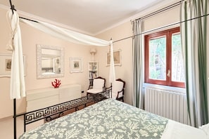 Ein Schlafzimmer mit einem Himmelbett, einem Fenster mit Vorhängen, einem Nachttisch, einem Spiegel und einer Heizung.