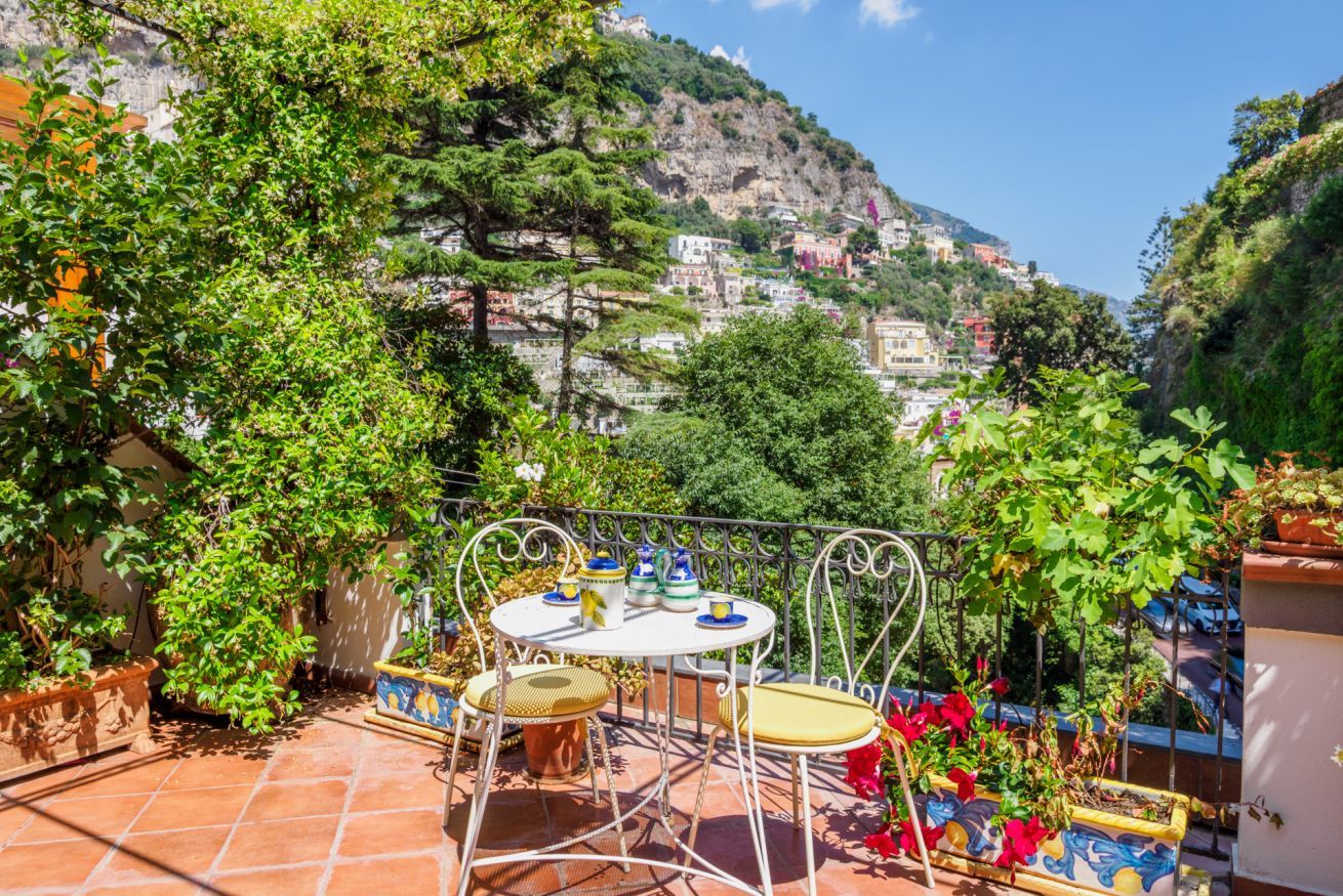 Una terraza con mesa y sillas, rodeada de plantas en macetas y con vistas a una colina con edificios.