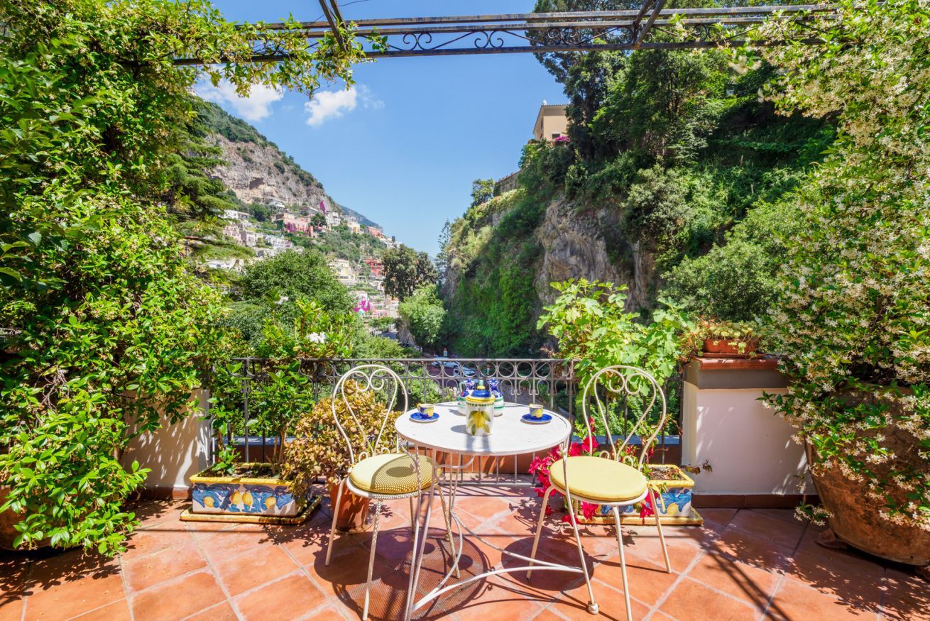 A terrace with a round table and chairs, surrounded by potted plants and a view of a mountainous landscape.