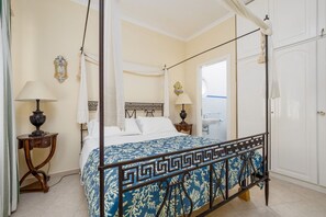 A four-poster bed with a geometric-patterned bedspread, two bedside tables with lamps, a wall-mounted decorative plate, and a white wardrobe.
