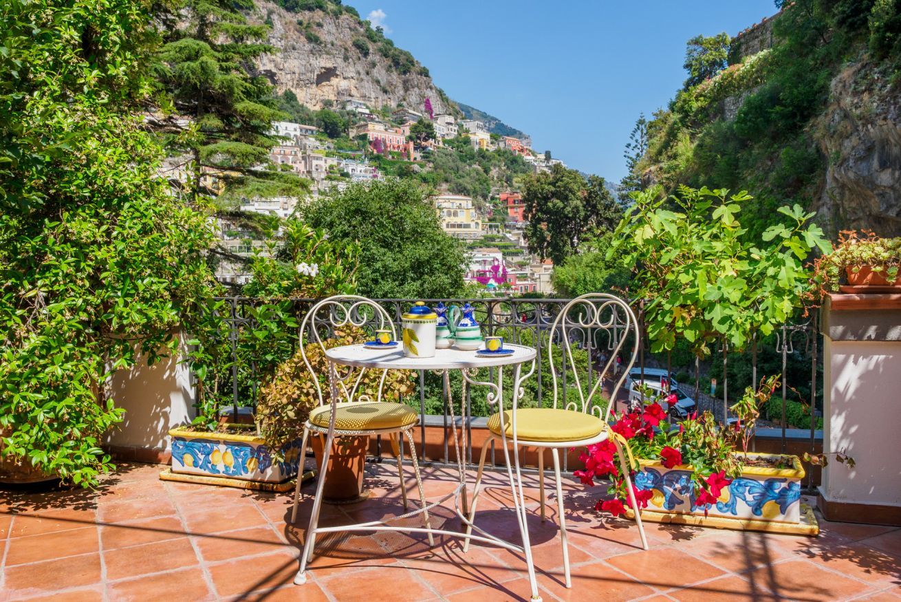 Una terraza con una mesa y sillas, rodeada de macetas con plantas y vistas a una colina con edificios.