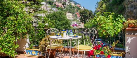 A terrace with a table and chairs, surrounded by potted plants and a view of a hillside with buildings.