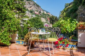 Terraço com mesa e cadeiras, rodeado por vasos de plantas e vista para uma colina com edifícios.