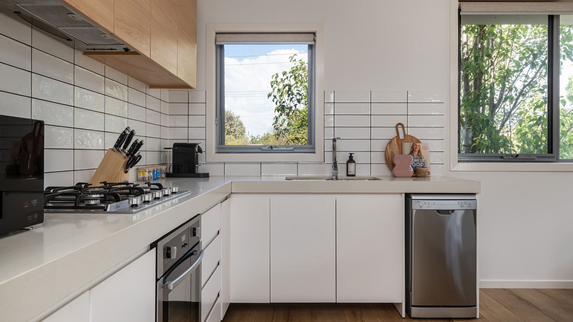 A modern kitchen with white cabinets, a stainless steel dishwasher, and a window with a view of trees.