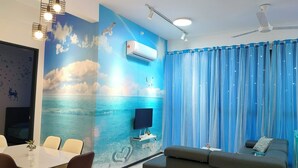 A room with a wall mural of a sea scene, a flat-screen TV, a ceiling fan, and a dining area with chairs and a table.