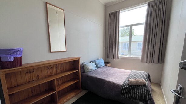 Una habitación pequeña con cama individual, un banco de madera, una ventana con cortinas y un espejo en la pared.