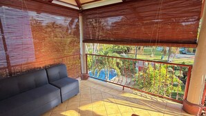 Un balcone coperto con un divano, una poltrona rossa e vista su una piscina e del verde.