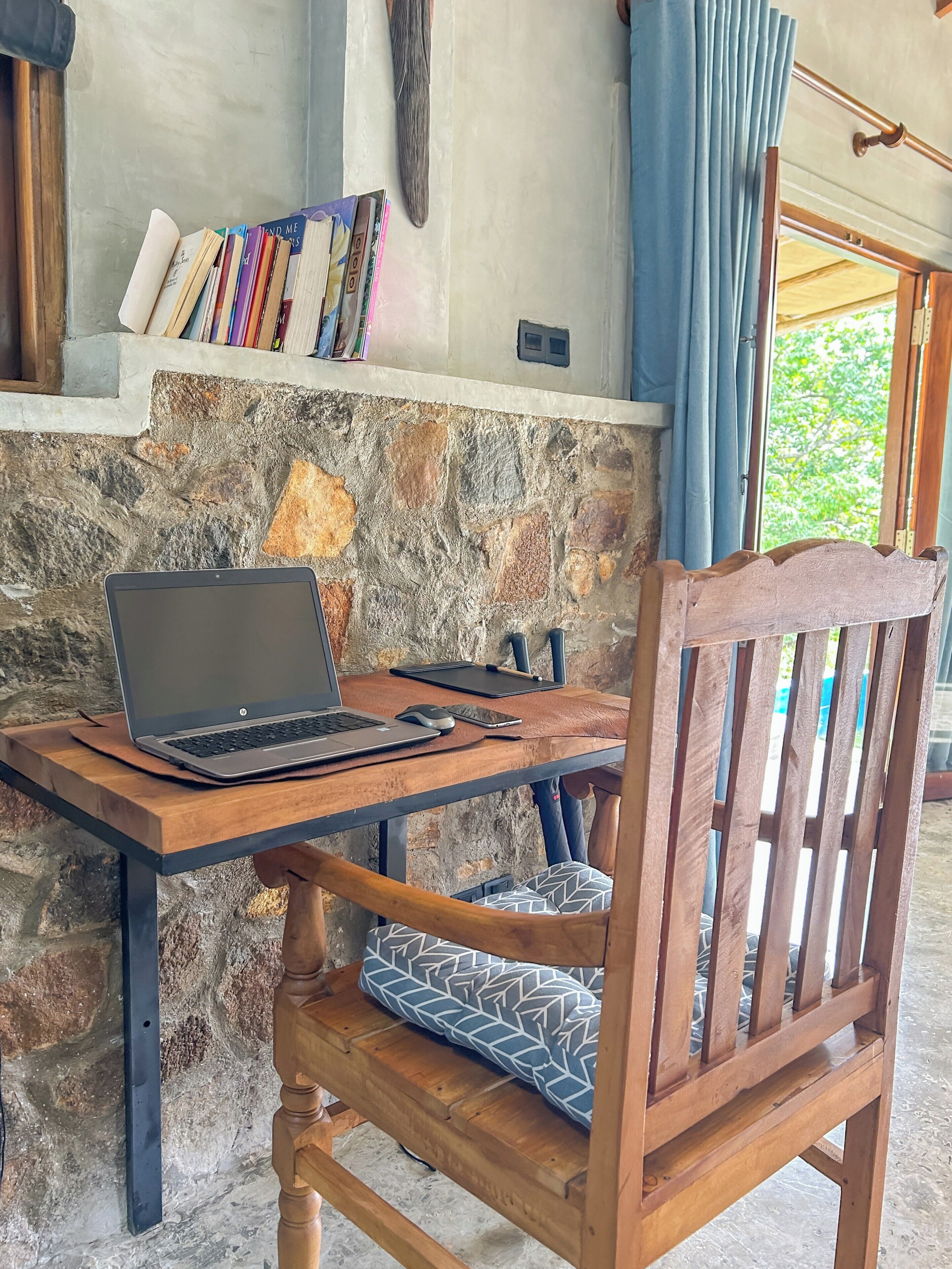 A wooden table with a laptop, a chair with patterned cushion, and books on a shelf against a stone wall.