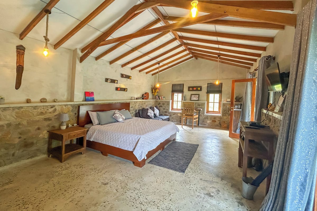 A bedroom with a bed, wooden beams, stone walls, a small table, and a chair.
