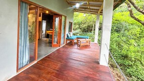 A covered outdoor area with a wooden floor, a seating area with a blue cushion, and a view of greenery. - Unseen Knuckles Eco Boutique  (Tawalanthanna)