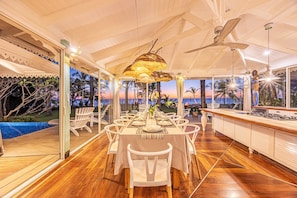 Un comedor con una larga mesa dispuesta para una comida, rodeada de sillas, con vista a palmeras y un atardecer.