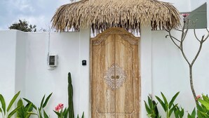 A white building with a wooden door, a thatched roof, and a small garden with plants and a cactus. - Casa Nimare (Ungasan)