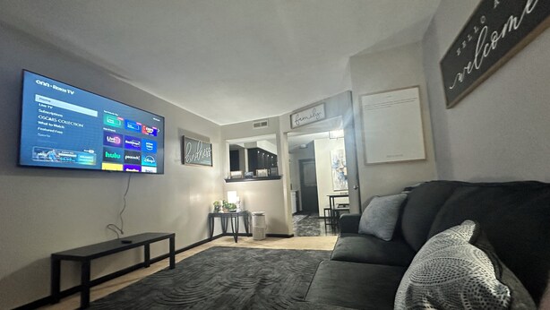 Een woonkamer met een flatscreen televisie waarop een streamingdienst te zien is, een donkergrijze bank met een gebreide sierkussen, een salontafel en een vloerkleed.