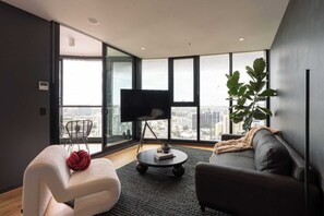 Una sala moderna con un sofá, un sillón blanco, una mesa de centro, un televisor sobre un mueble bajo y un ventanal con vista a la ciudad.