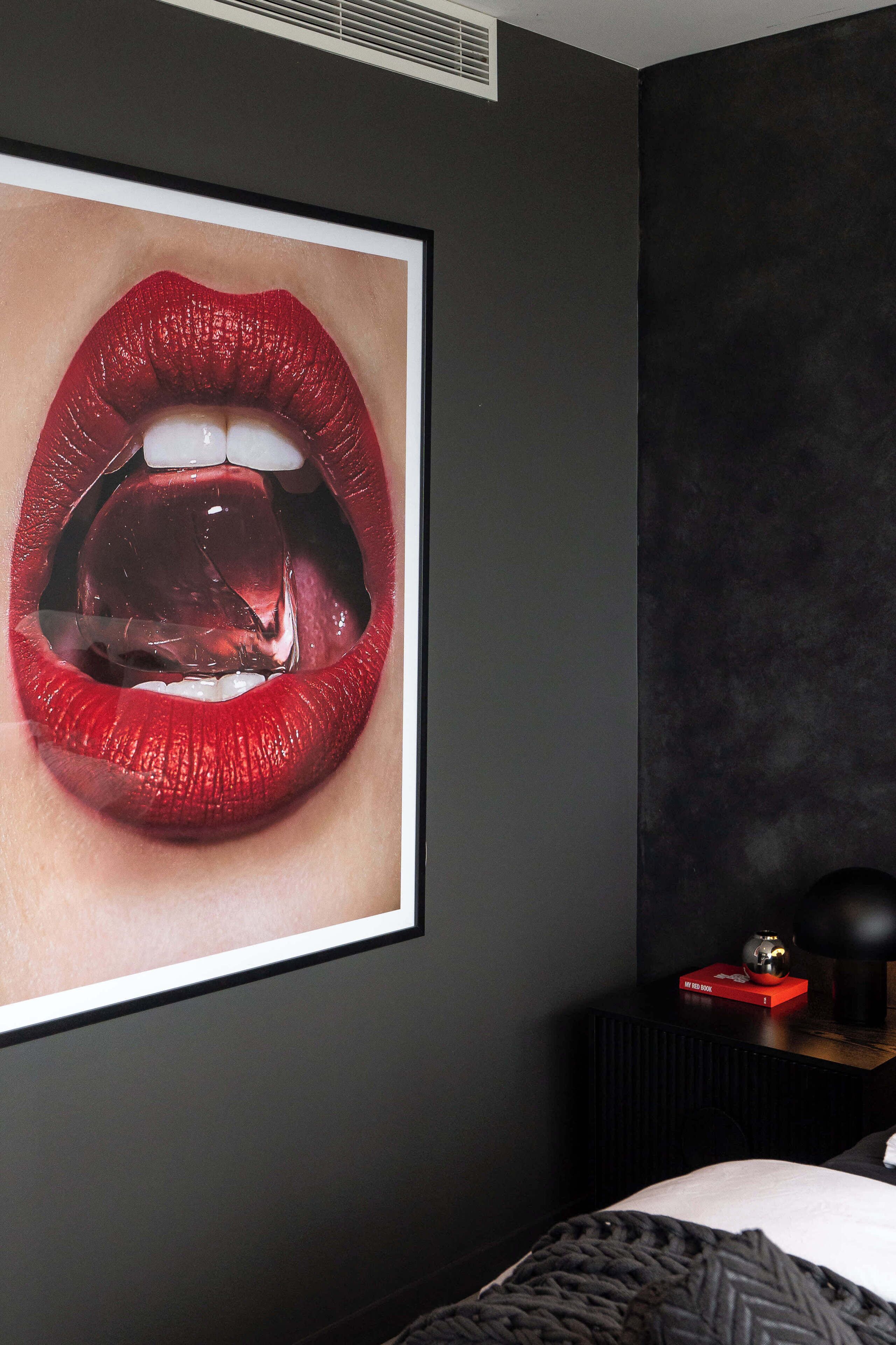 A framed artwork of lips on a wall, a bed with a patterned blanket, and a small table with a book and a spherical object.