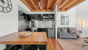 A modern kitchen with stainless steel appliances, a wooden dining table with bar stools, and a grey sofa in a living area. - 3-Bed Teneriffe Apartment - Pool | Gym | Parking (Teneriffe)