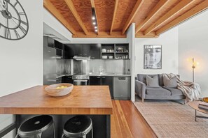 A modern kitchen with stainless steel appliances, a wooden dining table with bar stools, and a grey sofa in a living area. - 3-Bed Teneriffe Apartment - Pool | Gym | Parking (Teneriffe)