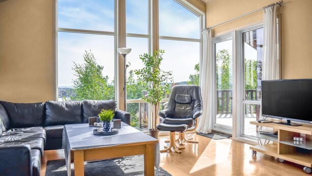 Un salon moderne comprenant un canapé, une table, un fauteuil, un téléviseur et de grandes fenêtres donnant sur un paysage verdoyant.