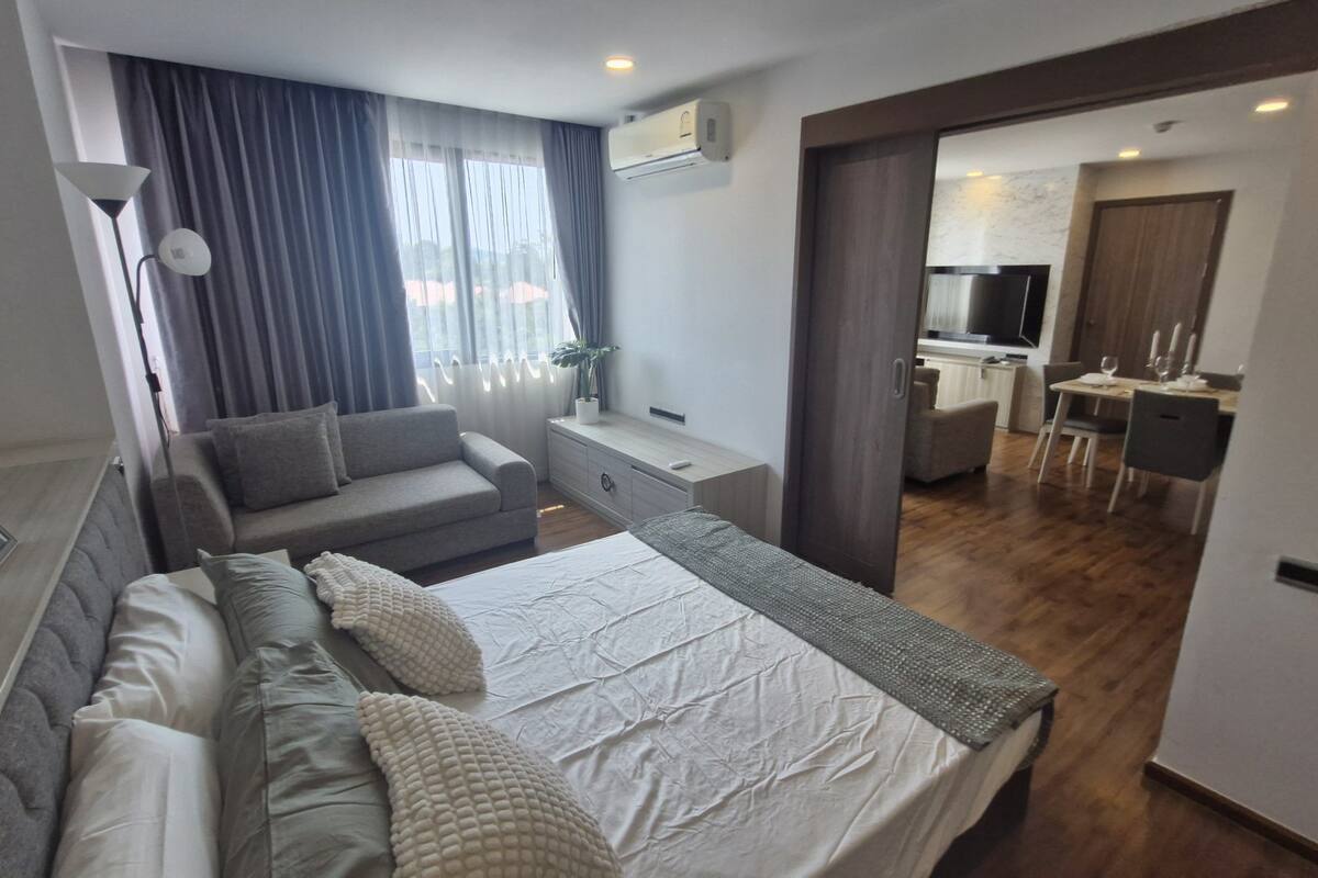 A modern hotel room with a bed, sofa, bedside tables, and a view of the dining area.