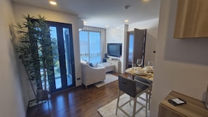 A modern living room with a sofa, a dining table set for two, a TV, and a large window with curtains. - Triple J Suite (Si Racha)