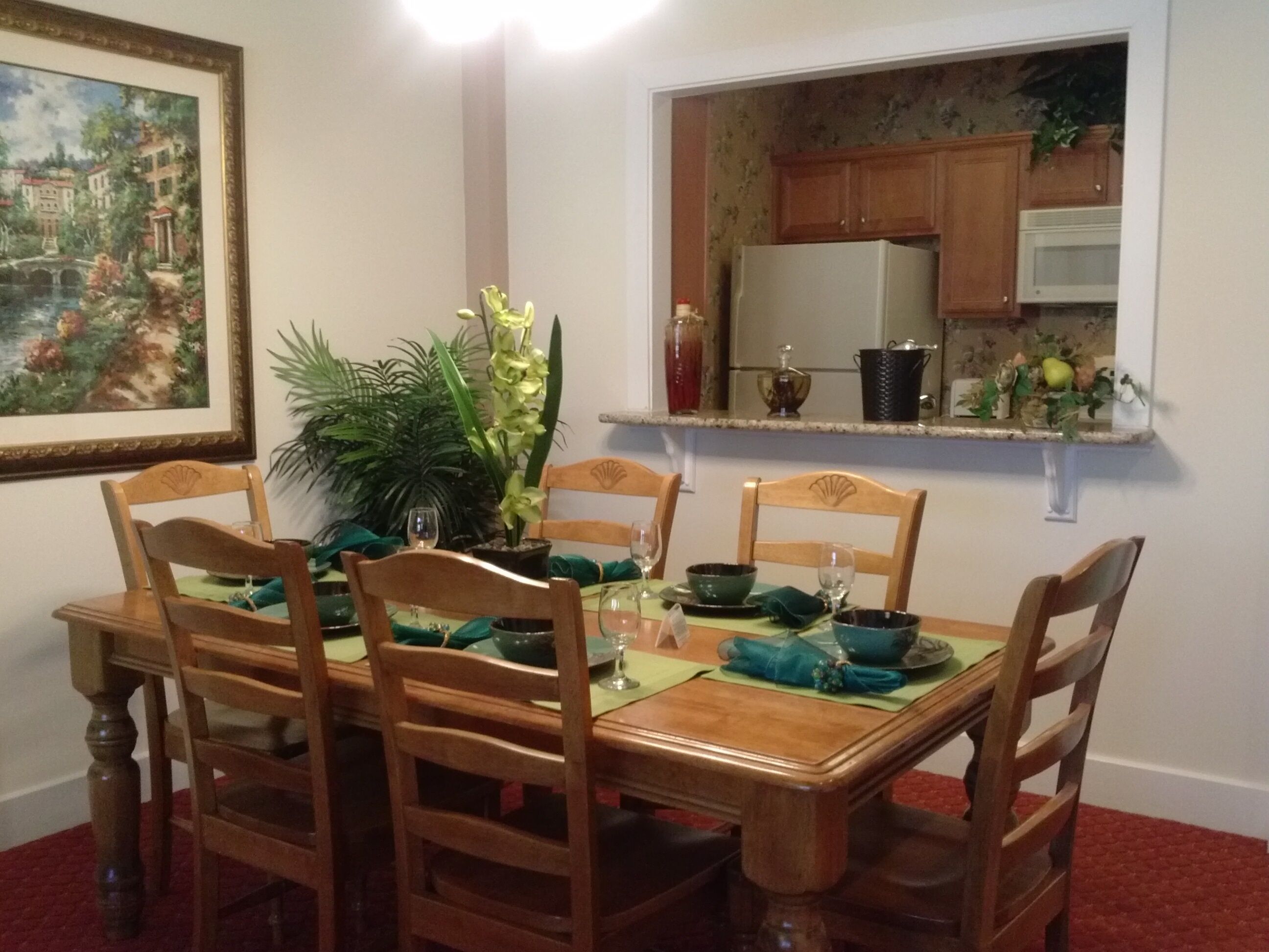 Une salle à manger avec une table en bois dressée pour quatre personnes, entourée de chaises. Un pot de fleurs est posé sur la table, et un tableau orne le mur.
