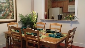Comedor con mesa de madera dispuesta para cuatro personas, rodeada de sillas. Hay una planta en maceta sobre la mesa y un cuadro en la pared.