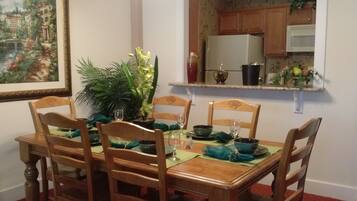 Une salle à manger avec une table en bois dressée pour quatre personnes, entourée de chaises. Un pot de fleurs est posé sur la table, et un tableau orne le mur.
