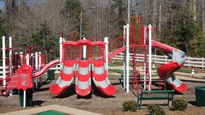 Un parque infantil con toboganes rojo y blanco, estructuras para escalar y una zona de juego verde.