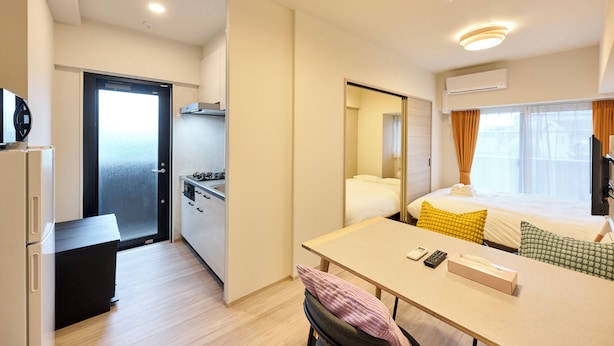 Ett kompakt hotellrum med ett litet pentry, en säng med kuddar, ett bord med en fjärrkontroll och ett fönster med gardiner.